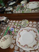 Photo shows Royal Albert plate with pink and yellow floral garlands and ribbon patterns, a small bowl with floral pattern, Denton China small floral basket, and Staffordshire floral bowl with minor chips all laid on a lace table cloth.