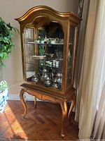 Full front view showing the gold painted cabinet with glass sides and displayed silverware and decorative items inside.