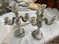 Photo showing both Dresden porcelain candle holders with multicolored floral decorations and gold accents, displayed on a lace tablecloth.