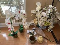 View of two faux orchid plants, one large arrangement in ceramic pot and one smaller plant in green pot, with extra stems and ceramic pot on floor near window.