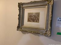 Framed needlepoint showing vintage-style house and trees from a side angle, highlighting the ornate silver and gold frame with cracks.