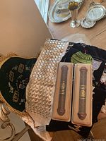 Two silver-tone trays and vase in background; pile of textiles including white satin scarf with silver embellishments and velvet hangings; two wooden mezuzah cases in plastic boxes in foreground