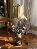 Full view of the lamp showing the fabric shade with tassels and floral metal and resin base on wooden floor near curtains.