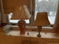 Two lamps on top of a white cabinet by a window showing the pink ceramic lamp with plastic-covered white shade and ribbed gold-green ceramic lamp with beige embroidered shade.