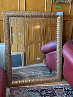 Full view of rectangular mirror showing the carved leaf motif wood frame with metallic gold finish, sitting on floor next to red chairs and against a wood paneled wall.