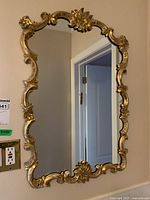 Front view of vertical rectangular mirror with ornate carved frame and gold finish mounted on beige wall.