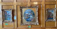 Three needlepoint art pieces framed with glass fronts hung on a wooden paneled wall, showing different designs including a wildlife scene and two coastal/village scenes.