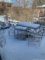 Snow-covered balcony showing large rectangular metal table with plexiglass top, metal chairs around, small mesh-shelf side table frame