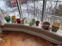 Wide view of assorted ceramic and wooden planters on a windowsill, various shapes and colors including a green small house-shaped planter, yellow and red pots, and a wicker basket.