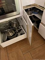 Photo of open kitchen cabinet drawers showing various stainless steel pots and pans with lids, along with metal strainers and kitchen tools.