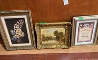 Three framed prints/paintings standing side by side against wooden wall, showing all items in the lot.