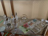 Wide view of assorted glass and crystal servingware including salt and pepper shakers, bowls, trays, and bottles on a glass shelf.