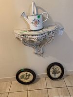 Front view of ceramic wall shelf with watering can on top, two oval framed florals below