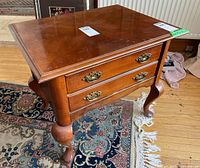 Front and side view of wooden side table showing two drawers with ornate metal handles and curved legs. The wood finish is medium brown with some wear.