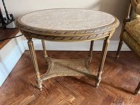 Full front view showing round beige marble top, carved gilt wood frame, four fluted legs and wicker lower shelf