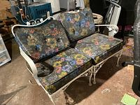 Three-quarter front view of white metal loveseat with floral vinyl cushions in basement setting