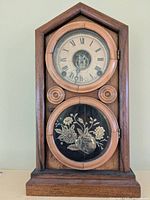 Front view of an antique wooden mantel clock showing two circular face windows: a clock dial with Roman numerals and an artistic floral glass panel below.
