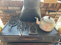 Four cast iron trivets and a metal kettle displayed on a cast iron stove top with brick background