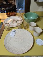 Full view of the Lenox wedding plate, mugs, Fire King bowls, Royal China plates, leaf bowl, cello bottle, gilded glasses and tea bag holder arranged on yellow surface.