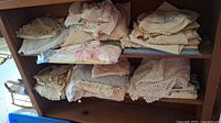 Two shelves full of assorted lace linens, doilies and folded linens stacked, showing various fabrics and colors mostly in beige, cream, and pink tones.