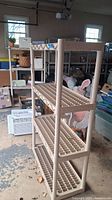 Beige plastic shelving unit with four grid shelves seen from the side in garage setting. Shelves have square lattice design.