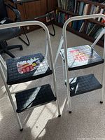 Two folding step stools with white metal frames and black textured steps, visible wear consistent with use, situated on carpet with wooden bookshelves in background.