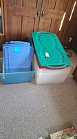 Nine assorted plastic storage bins and lids stacked on carpeted floor against wooden cabinet. Includes blue, clear with red rim, green lid bins and additional blue lids.