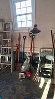 Wide view of yard tools and ladder including two shovels, three rakes, a red engine trimmer, and green hose reel with aluminum ladder to left.