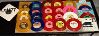 Photo showing all 23 colored vinyl 7-inch 45 RPM records laid out on a surface, showcasing various colors such as red, blue, orange, yellow, pink, and clear with white record sleeves in the background.
