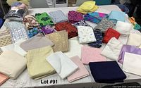 Overview of assorted folded fabrics and craft materials on a white table including patterned, solid, and decorative textiles in various colors.