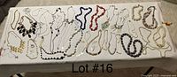 Wide image showing around 25 assorted costume jewelry necklaces laid out on a white surface, including faux pearls, chains, and colorful beaded styles.