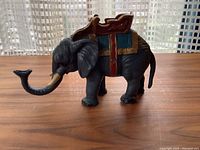 Side view of vintage cast iron elephant coin bank showing black body, detailed saddle in red and gold, curved trunk, and tail.