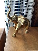 Front side view of a brass elephant figurine standing on a wooden surface in front of a window with patterned curtain, showing trunk raised and ears spread.