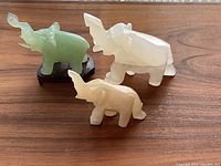 Three hand carved elephant ornaments displayed on wooden table: green jade elephant on wooden base, white and off-white stone elephants.