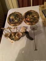 Assorted vintage decorative items on white surface including two ornate oval framed pictures with classical scenes, decorative scale, figurines, and wire skimmer.
