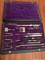 Opened vintage drawing and protractor tool set case showing multiple metal tools neatly arranged on purple velvet lining
