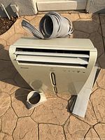 Front side view of the Sharp portable air conditioner with vent hose coiled on top and venting parts beside it on the ground outdoors.