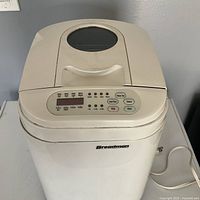 Top and front view of white Breadman bread maker showing control panel with buttons and digital display, and power cord on side.