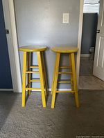Two yellow wooden stools against a wall showing front and side views.