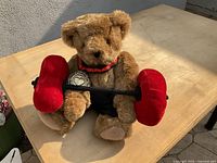 Brown teddy bear holding a plush barbell with red padded weights on a wooden table