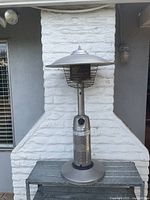 Full view of the stainless steel outdoor garden heater on the metal bench in front of a white textured brick wall.