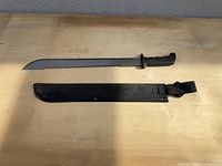 Photo of machete knife laid flat on a table next to its black fabric sheath with securing flap.
