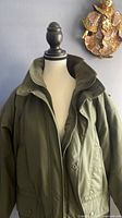 Olive green men's puffy jacket shown on mannequin with front zipper partially open and button flap visible, highlighting the collar and hood area.