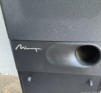 Close up of the Mirage logo and bass reflex port on a black speaker cabinet