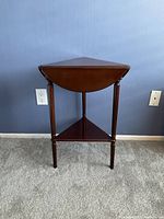 Front view of wooden side table showing semi-circular drop-leaf top and triangular lower shelf with turned legs.