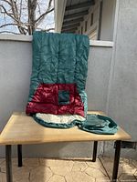 Two teal colored sleeping bags with red and cream interior laid out on table, each with matching teal carry bag visible.