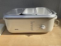 Side view of the electric slow cooker showing the dial with multiple cooking settings and three aluminum pans on top.