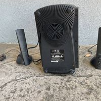 Back view of the black plastic subwoofer speaker showing circular vent and label details; left slim satellite speaker partially visible.