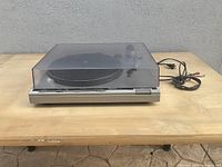 Side view of Technics SL-B3 turntable with dust cover and attached power cable and RCA cables on wooden table.