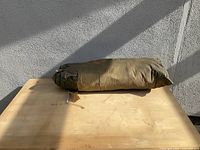 Green tent carry bag lying on wooden table against gray wall with sunlight casting shadows.